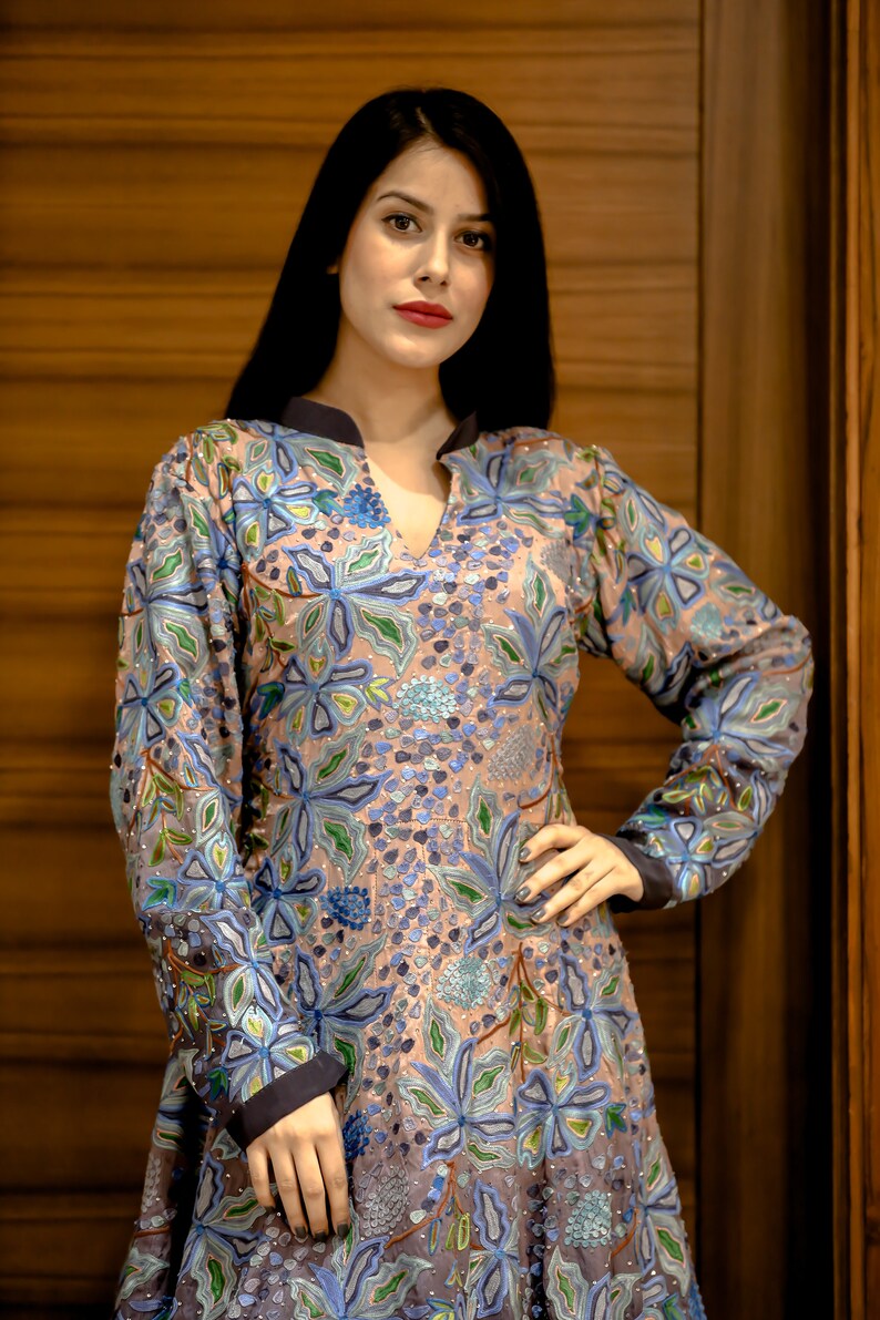 May include: A woman wearing a long-sleeved, light pink dress with blue and green floral embroidery. The dress has a V-neck and a fitted bodice.