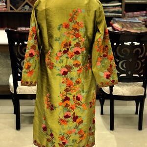 May include: A long-sleeved olive green tunic featuring intricate floral embroidery in shades of orange, pink, and deep red. The garment has a high collar and a straight silhouette, with the floral pattern extending from the neckline to the hem.