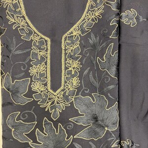 May include: A dark charcoal gray fabric garment featuring intricate embroidery of large leaves and vines. The embroidery is executed in a combination of black thread and metallic gold thread, which outlines the leaf patterns and the neckline.