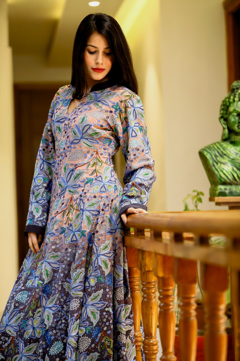 May include: A woman wearing a long, flowing dress with a floral pattern in shades of blue, green, and pink. The dress has a V-neckline and long sleeves. The woman is standing on a wooden staircase.