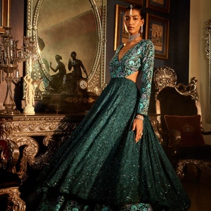 Pode incluir: Uma mulher vestindo um vestido verde-azulado com padr&atilde;o floral. O vestido tem um decote profundo e um corpete justo. A saia &eacute; cheia e fluida. A mulher est&aacute; em p&eacute; em um quarto com m&oacute;veis ornamentados e um espelho.