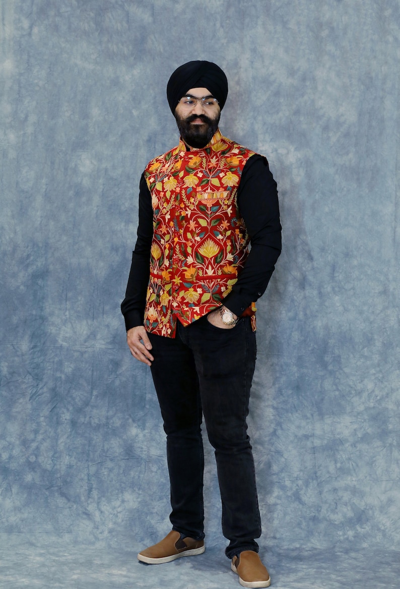 Op de afbeelding: Een man draagt een zwarte turban, een rood vest met bloemenprint, een zwart overhemd met lange mouwen, een zwarte jeans en bruine schoenen.
