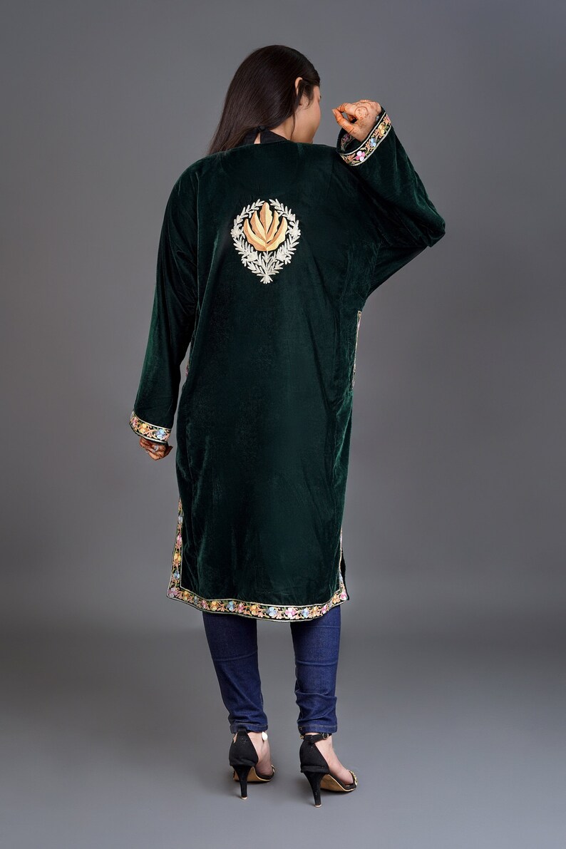 May include: A dark green velvet tunic featuring long sleeves and a high neckline. The back displays a large, central embroidered floral motif in gold and white, with colorful floral embroidery along the sleeve cuffs and the bottom hemline.