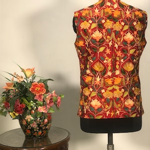 Op de afbeelding: Een rood vest zonder mouwen met een bloemenpatroon in tinten oranje, geel, groen en wit. Het vest heeft een mandarijnskraag en is gemaakt van een zachte, luxe stof.