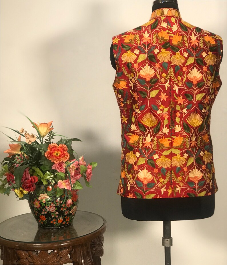 Op de afbeelding: Een rood vest zonder mouwen met een bloemenpatroon in tinten oranje, geel, groen en wit. Het vest heeft een mandarijnskraag en is gemaakt van een zachte, luxe stof.