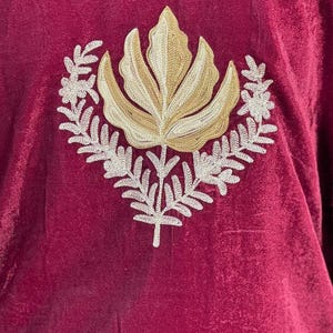 May include: A deep burgundy velvet fabric featuring a central embroidered design. The embroidery consists of a large, stylized golden-tan leaf shape positioned above a symmetrical arrangement of white, fern-like botanical branches.