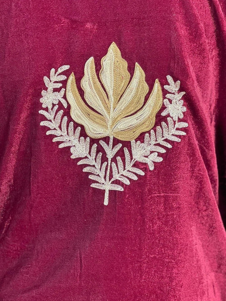 May include: A deep burgundy velvet fabric featuring a central embroidered design. The embroidery consists of a large, stylized golden-tan leaf shape positioned above a symmetrical arrangement of white, fern-like botanical branches.