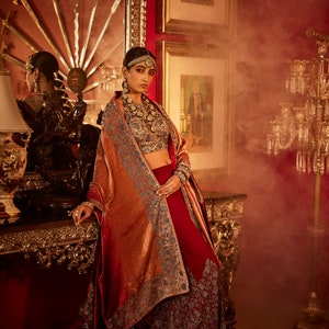 May include: A woman wearing a red and gold embroidered dress with a matching shawl. She is standing in a room with ornate furniture and a chandelier.