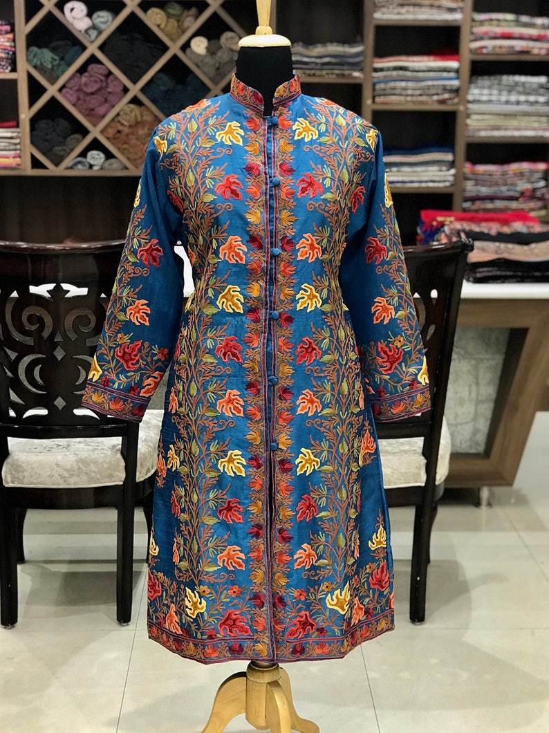 May include: A long, blue embroidered jacket with a mandarin collar and floral patterns. The jacket has a front closure with buttons and a long sleeve.