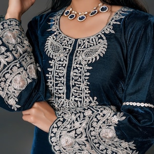 May include: A teal velvet dress with intricate silver embroidery on the bodice and sleeves. The dress features a round neckline and long sleeves. A gold necklace with blue gemstones complements the outfit, creating an elegant look.