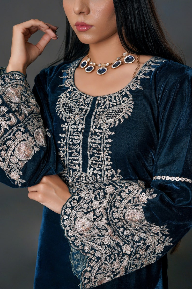May include: A teal velvet dress with intricate silver embroidery on the bodice and sleeves. The dress features a round neckline and long sleeves. A gold necklace with blue gemstones complements the outfit, creating an elegant look.