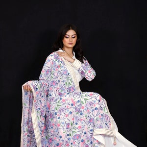 May include: A white embroidered dupatta with a floral pattern in shades of pink, blue, and green. The dupatta has a gold border and is draped over a white dress.