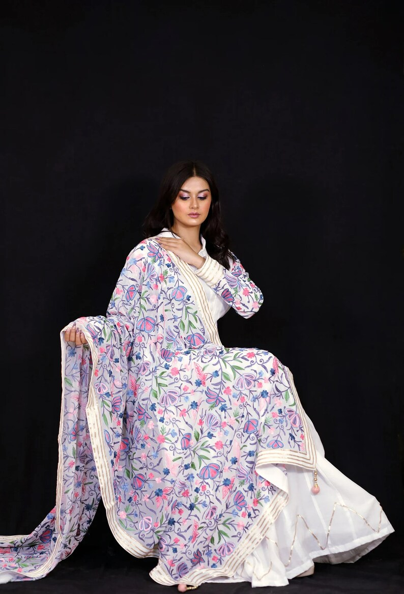 May include: A white embroidered dupatta with a floral pattern in shades of pink, blue, and green. The dupatta has a gold border and is draped over a white dress.
