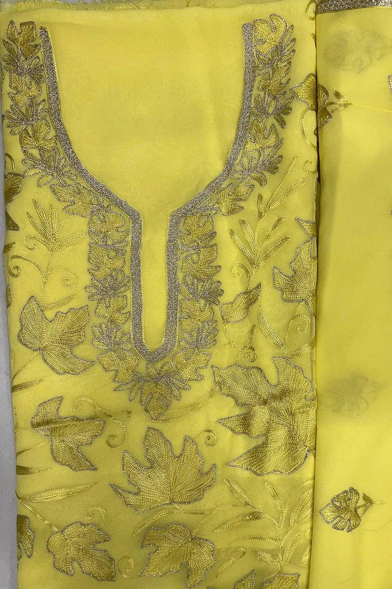 May include: A bright yellow fabric garment featuring intricate embroidery in a matching yellow thread. The design includes detailed leaf motifs scattered across the surface and a prominent, U-shaped neckline bordered by a metallic silver trim.