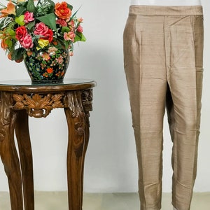 May include: Tan-colored pants with a subtle sheen, displayed on a mannequin. The pants have a tailored fit with a straight leg. A decorative wooden side table with carved details and a glass top is next to the pants. A vase of colorful flowers sits on the table.