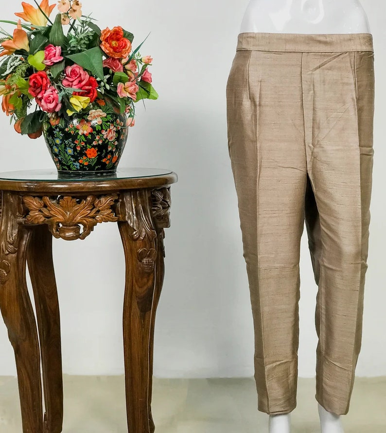 May include: Tan-colored pants with a subtle sheen, displayed on a mannequin. The pants have a tailored fit with a straight leg. A decorative wooden side table with carved details and a glass top is next to the pants. A vase of colorful flowers sits on the table.