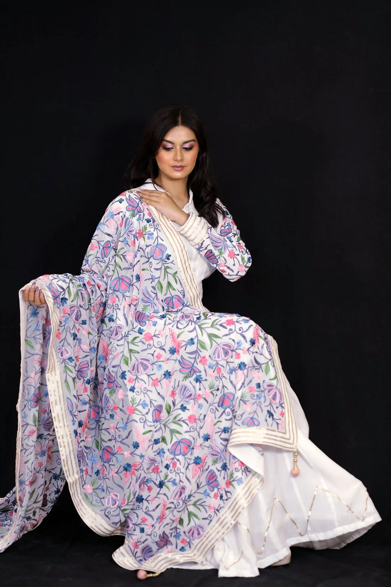 May include: A white embroidered dupatta with a floral pattern in shades of pink, blue, and green. The dupatta has a gold and white border.