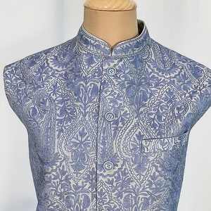 Puede incluir: Un chaleco azul claro con fondo blanco y estampado paisley. El chaleco tiene cuello mandar&iacute;n y cierre de botones.