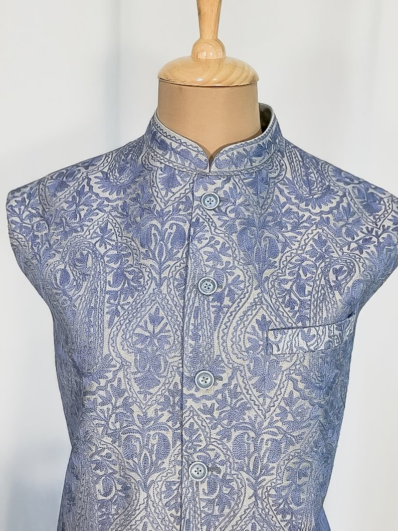 Puede incluir: Un chaleco azul claro con fondo blanco y estampado paisley. El chaleco tiene cuello mandar&iacute;n y cierre de botones.