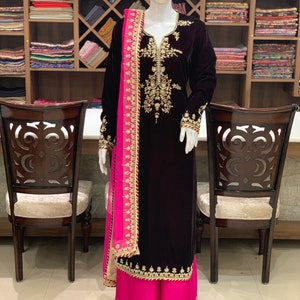May include: A maroon velvet kurta with gold embroidery and a pink dupatta. The kurta has a V-neckline and long sleeves. The dupatta is sheer and has a gold border. The kurta is paired with pink palazzo trousers.