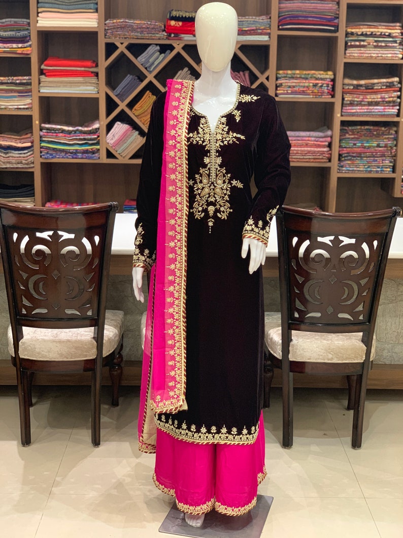 May include: A maroon velvet kurta with gold embroidery and a pink dupatta. The kurta has a V-neckline and long sleeves. The dupatta is sheer and has a gold border. The kurta is paired with pink palazzo trousers.