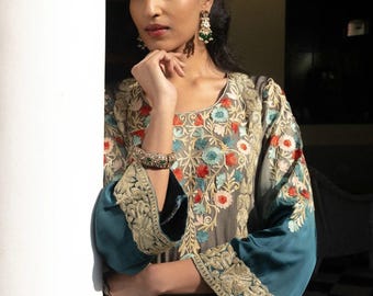 May include: A woman wearing a gray dress with floral embroidery and teal sleeves. The dress features intricate gold detailing on the sleeves and neckline. She is also wearing green earrings and a gold bracelet.