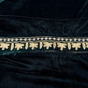 May include: A dark green velvet fabric with a gold and silver embroidered trim. The trim features a repeating pattern of small leaves.