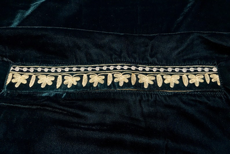 May include: A dark green velvet fabric with a gold and silver embroidered trim. The trim features a repeating pattern of small leaves.