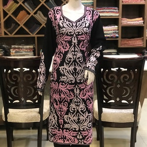 May include: A long-sleeved black tunic featuring a V-neckline and intricate, symmetrical embroidery in shades of light pink and white. The floral-inspired pattern covers the front and sleeves of the garment, which is displayed on a mannequin.