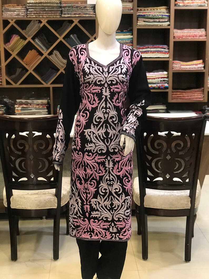 May include: A long-sleeved black tunic featuring a V-neckline and intricate, symmetrical embroidery in shades of light pink and white. The floral-inspired pattern covers the front and sleeves of the garment, which is displayed on a mannequin.
