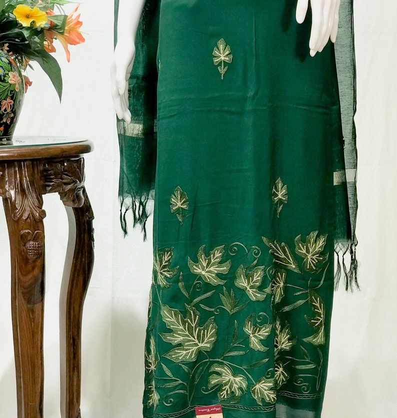 May include: A dark green dress with a leaf pattern embroidered on the bottom hem. The dress is made of a soft, flowing fabric and has a simple, elegant design.