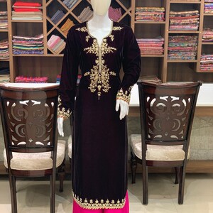 May include: A long, dark purple velvet kurta with gold embroidery. The kurta has a V-neckline and long sleeves. The bottom of the kurta is trimmed with gold embroidery. The kurta is paired with a pink palazzo pant.