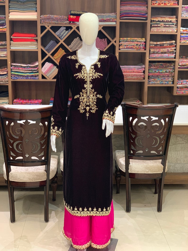 May include: A long, dark purple velvet kurta with gold embroidery. The kurta has a V-neckline and long sleeves. The bottom of the kurta is trimmed with gold embroidery. The kurta is paired with a pink palazzo pant.