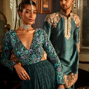 Pode incluir: Um casal em trajes tradicionais indianos. A mulher est&aacute; vestindo um vestido verde-azulado com bordados florais e um colar de prata. O homem est&aacute; vestindo uma kurta verde-azulada com bordados dourados.