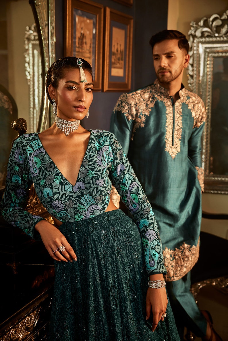 Pode incluir: Um casal em trajes tradicionais indianos. A mulher est&aacute; vestindo um vestido verde-azulado com bordados florais e um colar de prata. O homem est&aacute; vestindo uma kurta verde-azulada com bordados dourados.