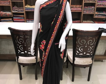 May include: A black saree featuring an intricate floral border embroidered with vibrant orange, pink, and green threads. The fabric drapes from the shoulder across the torso, showcasing a detailed pattern along the edges and a solid black base.