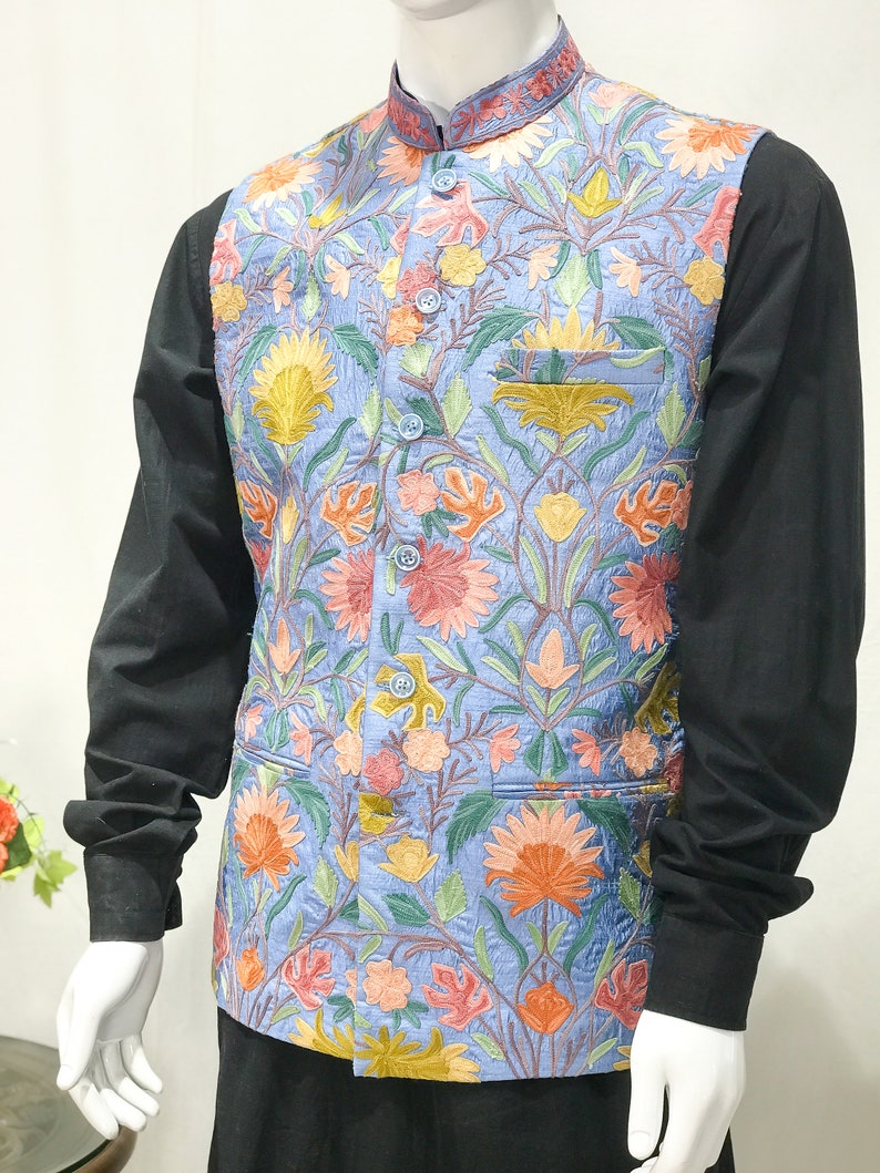 May include: A light blue vest with a floral pattern embroidered in shades of pink, yellow, orange, and green. The vest has a mandarin collar and a button closure.