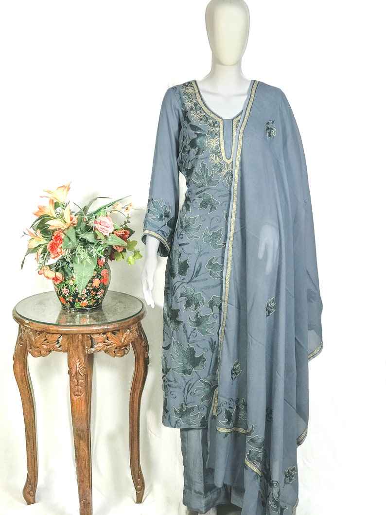 May include: A slate blue long-sleeved tunic and matching trousers set featuring dark green leaf embroidery throughout. The ensemble includes a coordinating sheer slate blue dupatta scarf with gold trim and matching leaf embroidery along the edges.