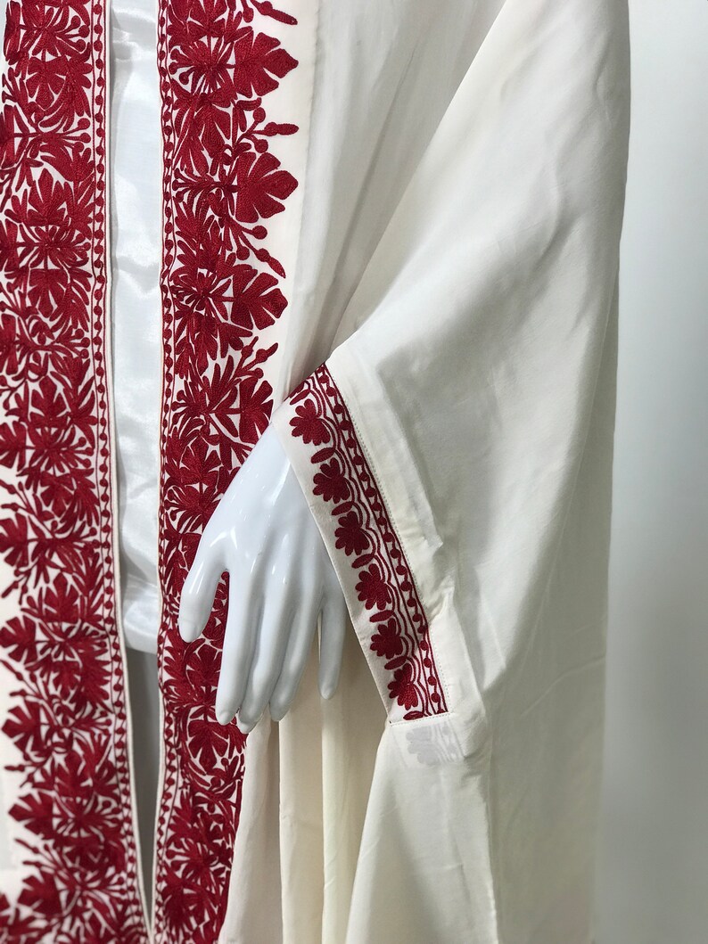 May include: A white cotton robe with red embroidered floral designs along the edges. The robe has a long, flowing silhouette and a wide, open neckline.