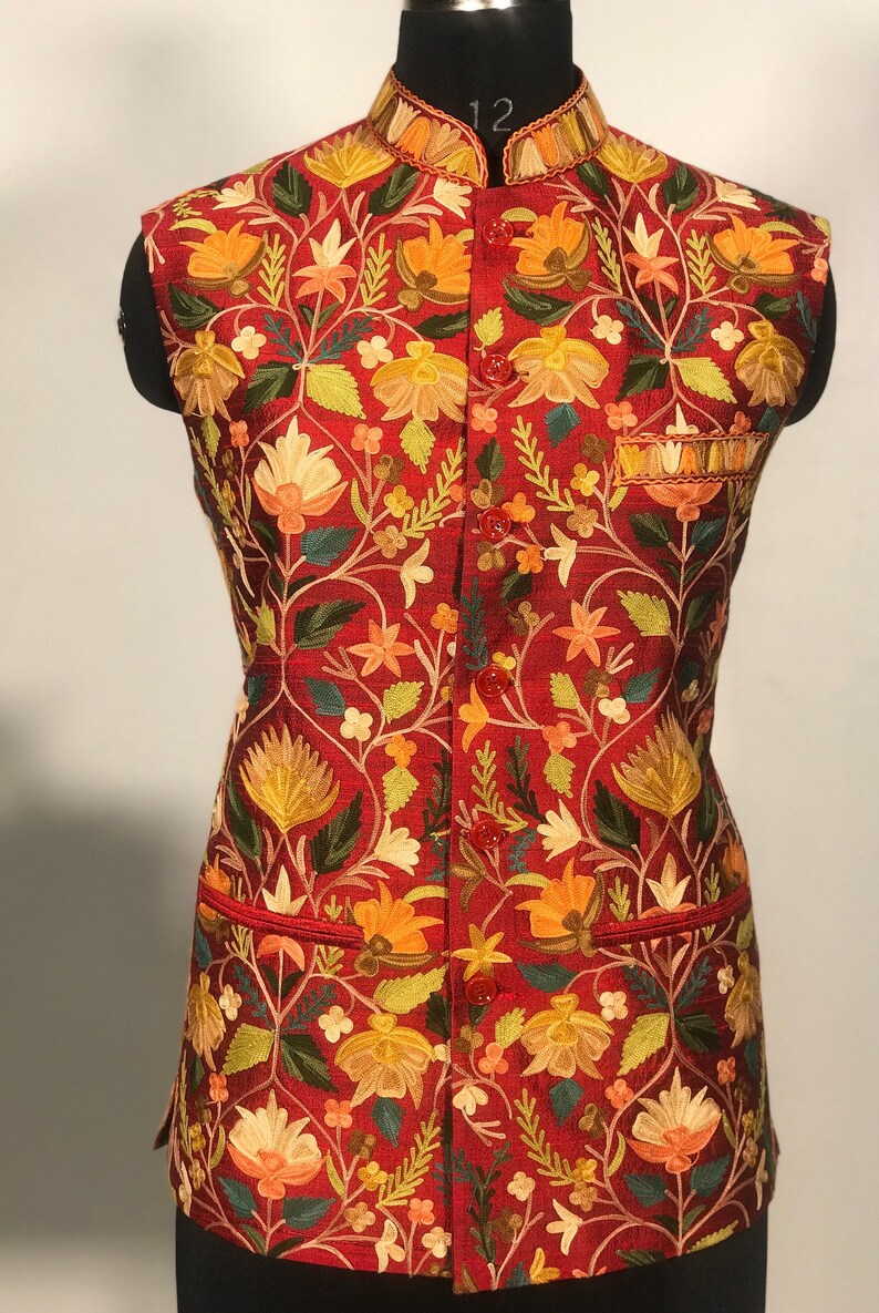 Op de afbeelding: Een rood, mouwloos vest met een bloemenpatroon geborduurd in tinten geel, oranje, groen en wit. Het vest heeft een mandarijnskraag en een knoopsluiting.