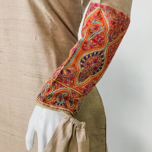 May include: A tan-colored garment with intricately embroidered sleeves. The sleeves feature a vibrant floral pattern in shades of orange, red, blue, and green. The cuff is gathered, and the fabric has a subtle sheen.