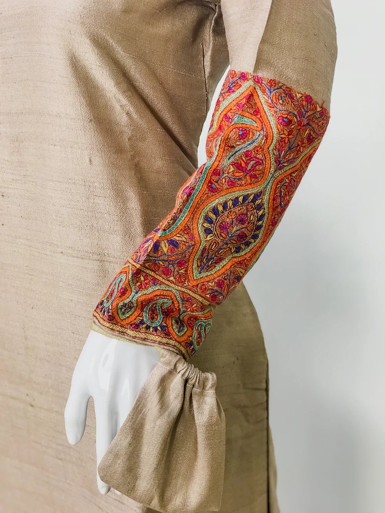 May include: A tan-colored garment with intricately embroidered sleeves. The sleeves feature a vibrant floral pattern in shades of orange, red, blue, and green. The cuff is gathered, and the fabric has a subtle sheen.