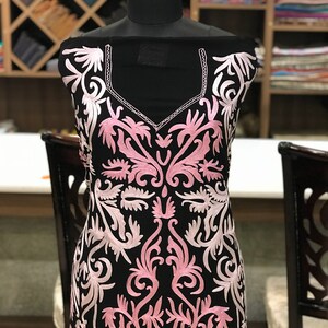 May include: A black tunic featuring a V-neckline and intricate light pink floral embroidery that covers the front. The garment is displayed on a mannequin, showcasing the detailed needlework and the contrast between the dark fabric and the soft pink thread.