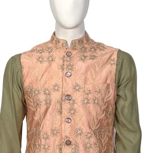 Op de afbeelding: Een perzikkleurig vest met een bloemenpatroon en knoopsluiting. Het vest heeft een zak aan de linkerkant en wordt gedragen over een groen shirt.