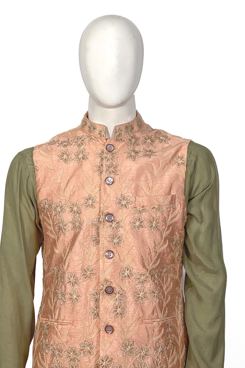 Op de afbeelding: Een perzikkleurig vest met een bloemenpatroon en knoopsluiting. Het vest heeft een zak aan de linkerkant en wordt gedragen over een groen shirt.
