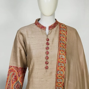 May include: A beige silk kurta suit with intricate embroidery. The mandarin collar and cuffs feature detailed floral patterns in red, orange, and blue. The suit has a row of decorative buttons down the front and a matching scarf.
