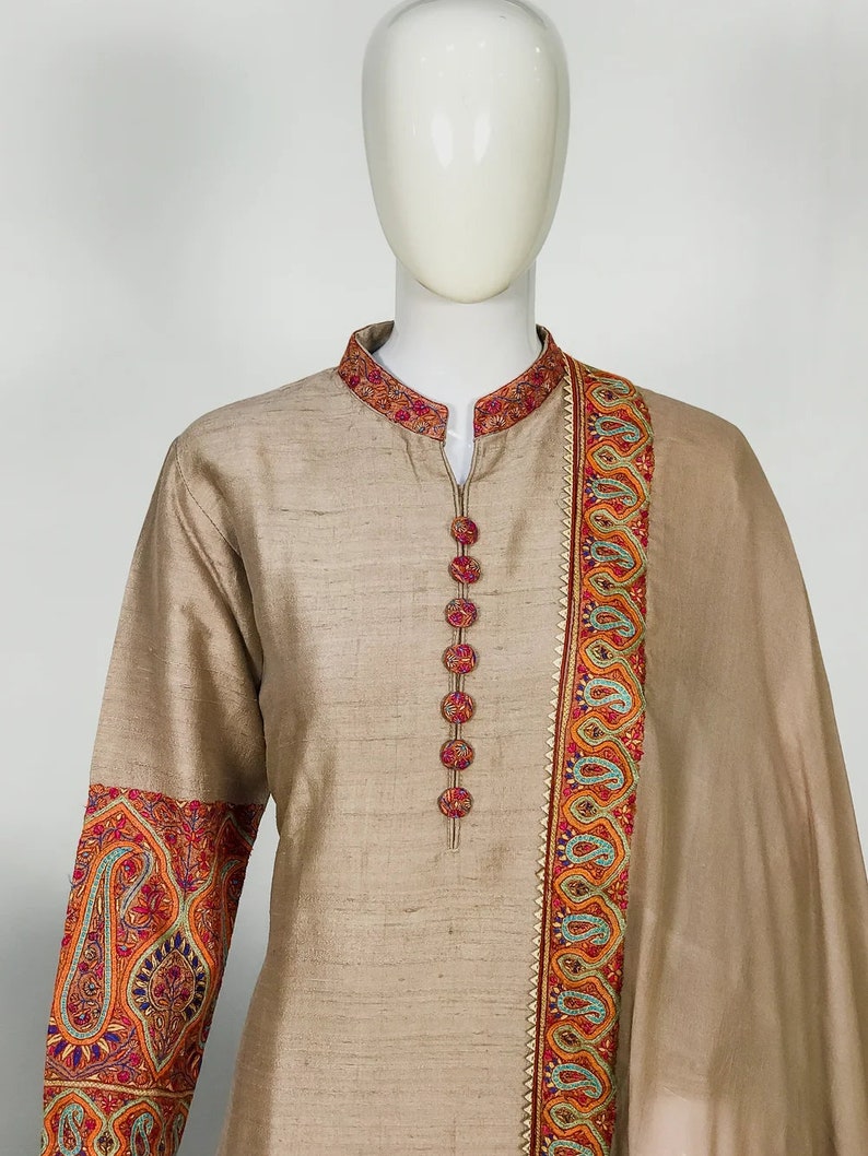 May include: A beige silk kurta suit with intricate embroidery. The mandarin collar and cuffs feature detailed floral patterns in red, orange, and blue. The suit has a row of decorative buttons down the front and a matching scarf.