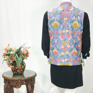 May include: A light blue vest with a floral embroidery pattern. The vest has a mandarin collar and a button closure. The vest is worn over a black shirt and white pants.