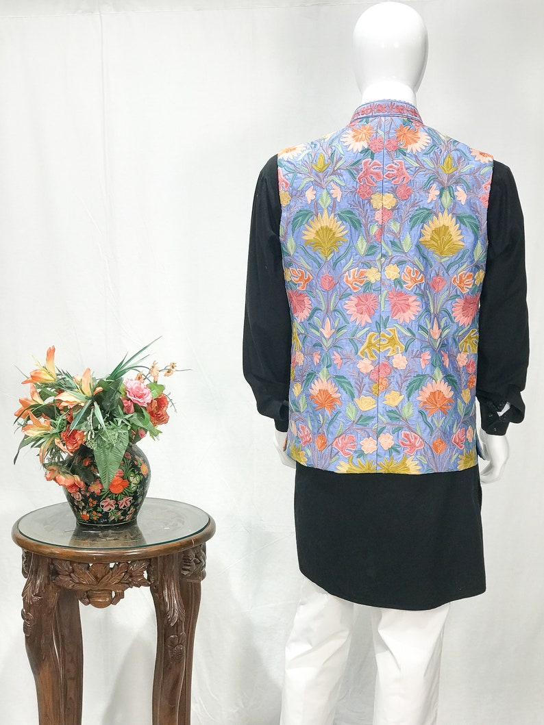 May include: A light blue vest with a floral embroidery pattern. The vest has a mandarin collar and a button closure. The vest is worn over a black shirt and white pants.