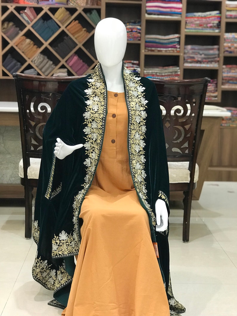 May include: A dark green velvet shawl with intricate white and gold embroidery. The shawl is draped over a mannequin wearing a long, orange dress.